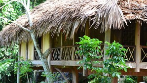 Une construction au toit de chaume, dotée d’un balcon en bois, entourée d’une végétation luxuriante.