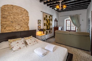Un dormitorio con pared de piedra, cama con almohadas estampadas, sofá, lámpara y vistas al exterior por una ventana.