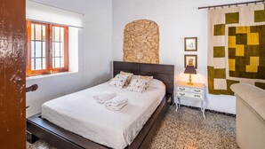Un dormitorio con cama, pared de piedra, ventana, mesita de noche y colcha.
