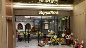 Entrada do PappaRich com interior moderno, portas de vidro e diversas flores expostas do lado de fora.
