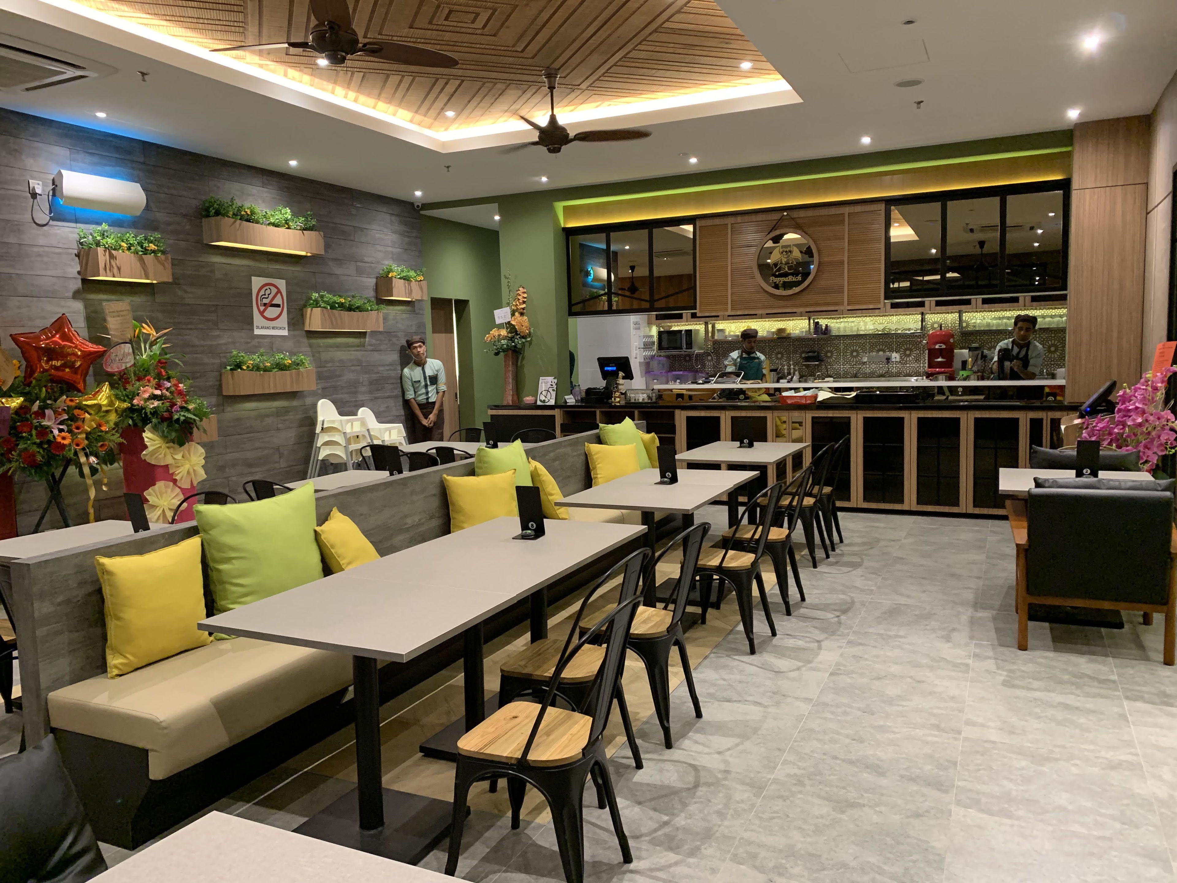 Un restaurante moderno con un comedor que incluye mesas largas y asientos acolchados. Se ve una zona de cocina al fondo.