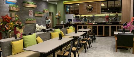 Un restaurante moderno con un comedor que incluye mesas largas y asientos acolchados. Se ve una zona de cocina al fondo.