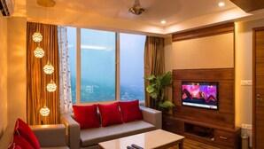A modern living room with a sofa, red cushions, a TV on a wooden stand, and a view of a cityscape through large windows. - Hotel Inclover (Dharamshala)