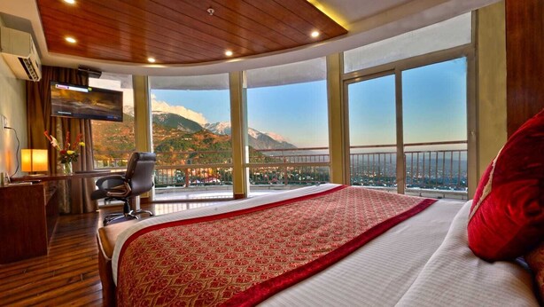 A hotel room with a large bed, a desk with a chair, and a television. The room has large windows with a mountain view. - Hotel Inclover (Dharamshala)