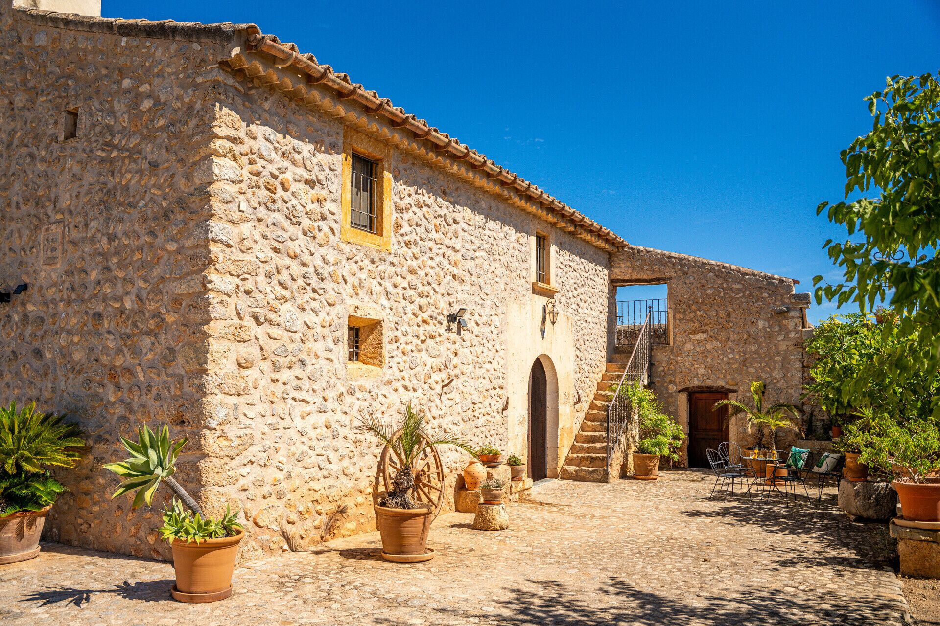Un bâtiment en pierre avec un toit en tuiles, une porte en bois et un escalier menant à un deuxième étage. On y trouve des plantes en pot et des sièges d’extérieur.