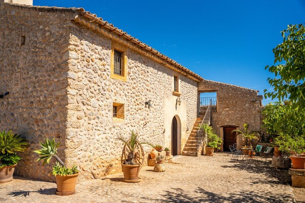 A stone building with a tiled roof, a wooden door, and a staircase leading to a second level. There are potted plants and outdoor seating. - Mallorcan finca Ses Planes with pool (Buger)