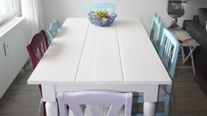 Zona pranzo con tavolo bianco, sedie blu, una ciotola di frutta e una lampada a sospensione.