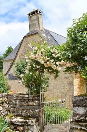 A stone building with a chimney, a garden with white flowers, and a metal gate.