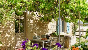 Un bâtiment en pierre avec une table et des chaises placées sous une pergola couverte de plantes grimpantes.