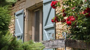 Un bâtiment en pierre avec deux fenêtres, dont une avec un volet, et une clôture en fil de fer ornée de roses.