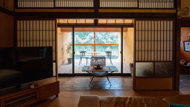 Ein traditioneller japanischer Raum mit Tatami-Boden, einem niedrigen Holztisch mit Stühlen, einem Fernseher auf einem Holzständer und Schiebetüren zu einem Außenbereich.