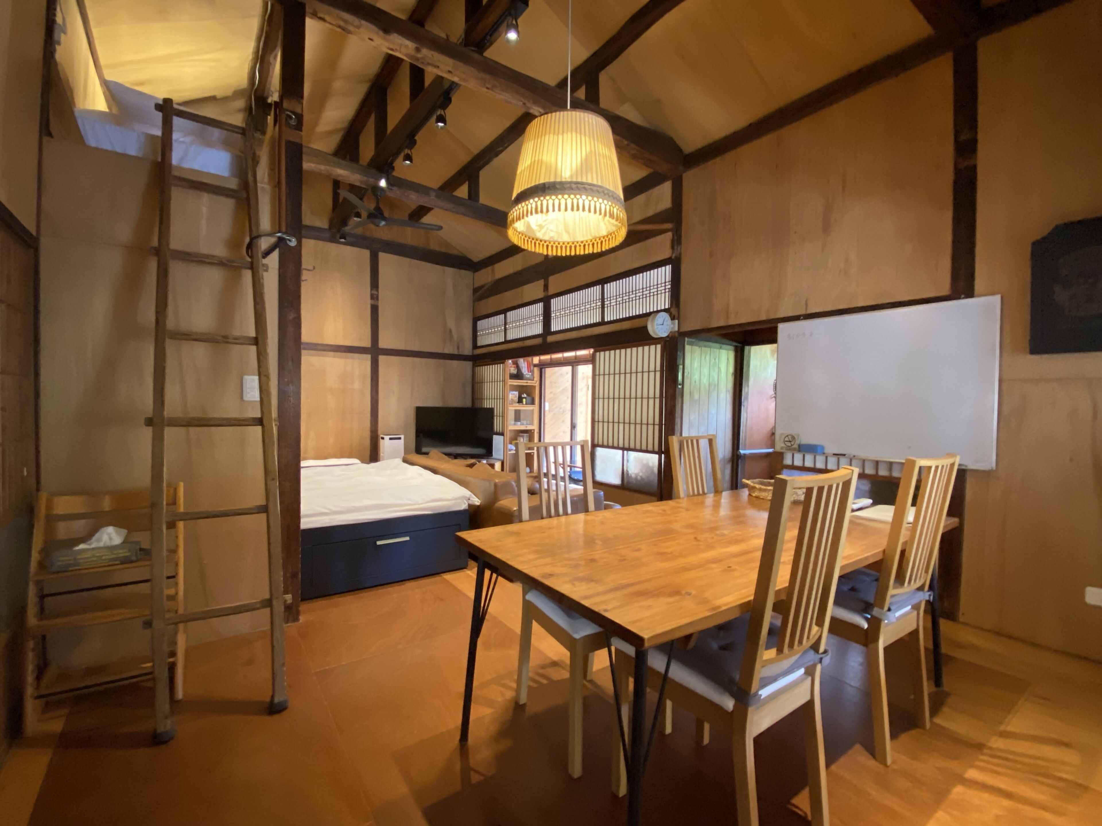 A traditional Japanese-style room with tatami flooring, a wooden dining table with chairs, a ladder, a bed, a television, and a whiteboard.