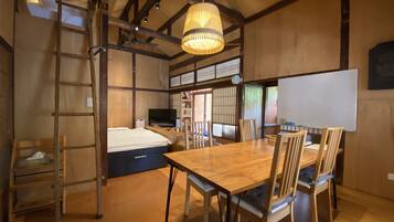 A traditional Japanese-style room with tatami flooring, a wooden dining table with chairs, a ladder, a bed, a television, and a whiteboard.