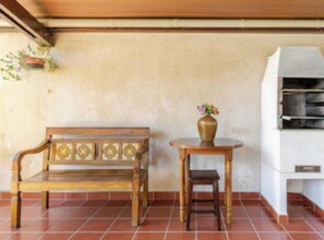 Un banco de madera y una mesa pequeña con un florero, frente a una pared lisa con suelo de baldosas.