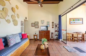 A living room with a sofa, coffee table, and a television. There are wall decorations and a dining area visible.