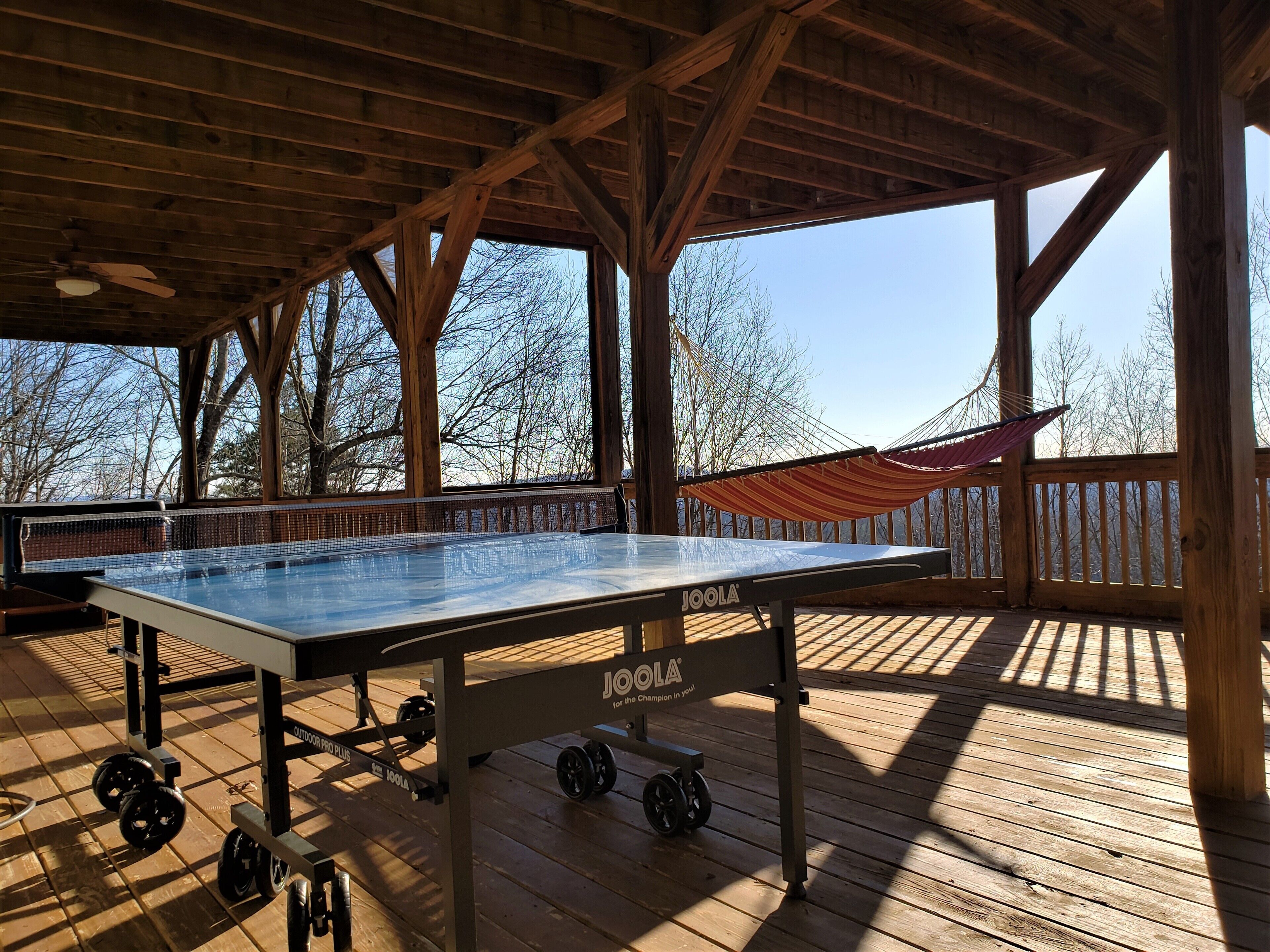 An outdoor ping pong table with wheels, a hammock, and a wooden deck with a view of trees.