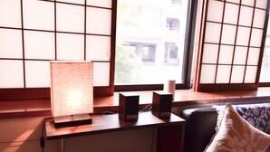 Een kamer met een bureau, een lamp, speakers en een raam met uitzicht op gebouwen.