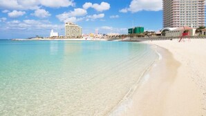Een strand met helder turquoise water, een zandstrand en een hoog hotel op de achtergrond.
