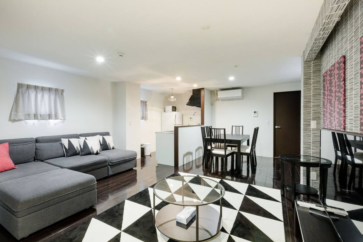 A modern living room with a grey sofa, a glass coffee table, and a dining area with a black and white checkered floor.