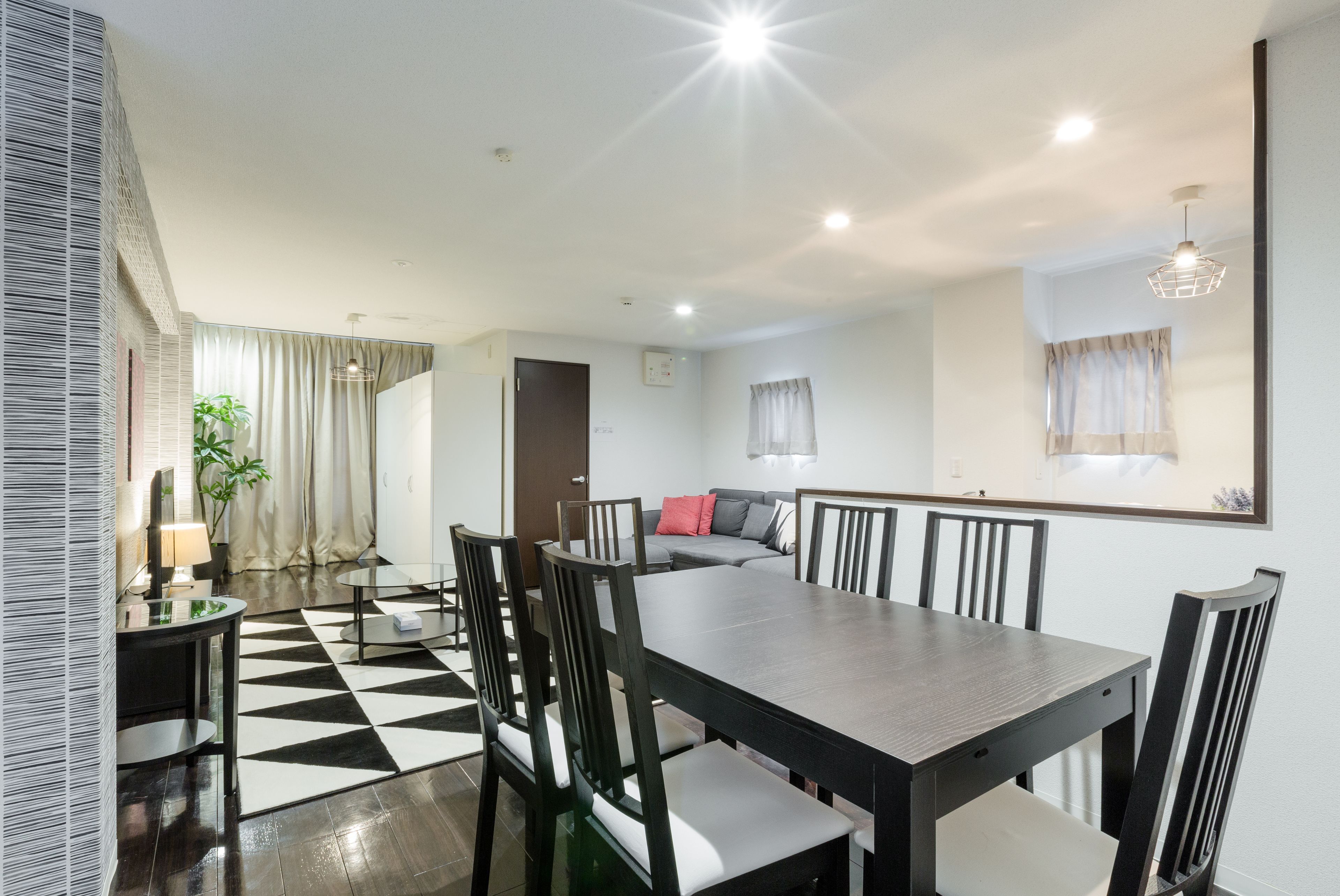 A dining area with a large rectangular table, black chairs, and a black and white checkered floor. There is a sofa with cushions in the background, a plant, and a window with curtains.