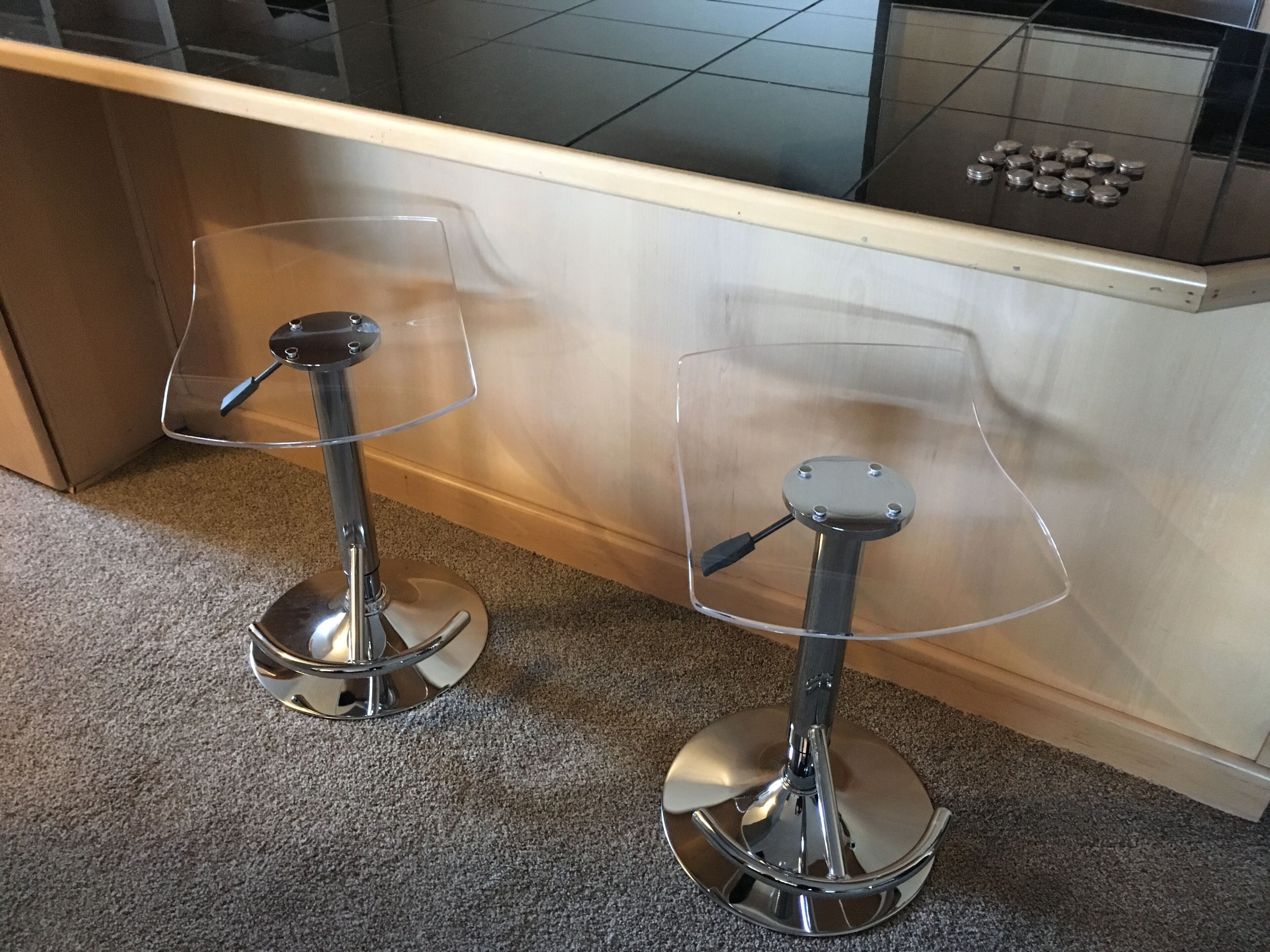 Zwei Barhocker mit durchsichtigen Sitzflächen und Chromfüßen auf einem Teppichboden nahe einer Holztresen.