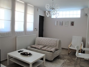 A modern living room with a sofa, a white table, and a chair. There are windows with blinds, a radiator, and a ceiling light fixture.