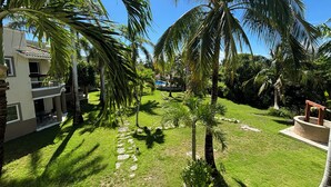 A well-maintained garden with palm trees, a clear blue sky, and a building with balconies. - Charming 2 Bedrooms Villa, 1 minute to Private Beach, Pool, Chef, Wi-Fi, Staff. (PLAYA DEL SECRETO)