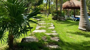 A tropical garden pathway with palm trees, a thatched-roof structure, and a pool area. - Charming 2 Bedrooms Villa, 1 minute to Private Beach, Pool, Chef, Wi-Fi, Staff. (PLAYA DEL SECRETO)