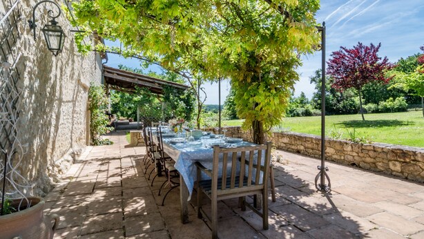 Ein Essbereich im Freien mit Tisch unter einer Pergola, Steinmauern und Blick auf eine grüne Landschaft.