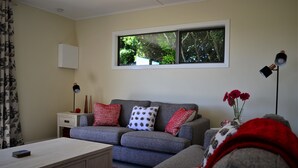Een woonkamer met een grijze bank, een witte bijzettafel, een rode en witte gebreide plaid en een raam met uitzicht op bomen.