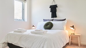 Un lit bien fait, avec du linge de lit blanc, deux lampes de chevet et une décoration en macramé suspendue au-dessus.