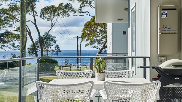 Un balcon avec des fauteuils en osier blanc, un barbecue et une vue sur la mer.