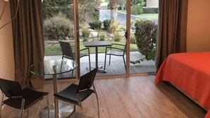Ein Hotelzimmer mit Glastür zu einer Terrasse, auf der ein Tisch und Stühle stehen. Ein Bett mit roter Bettdecke und eine Klimaanlage an der Wand sind ebenfalls zu sehen.