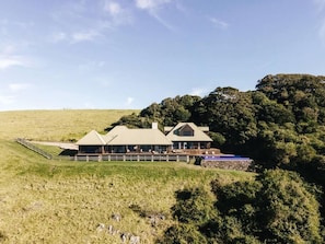 A large, modern building with a flat roof and a covered porch, surrounded by a grassy field and dense trees.