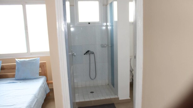 Quarto pequeno e luminoso com chuveiro, uma cama com colcha azul e uma janela.