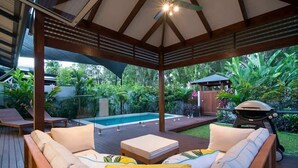 A covered outdoor seating area with a pool, wooden deck, and lush greenery.
