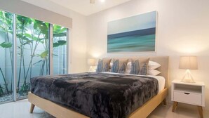 A modern bedroom with a large bed, a view of greenery through glass doors, and a painting of a beach scene.