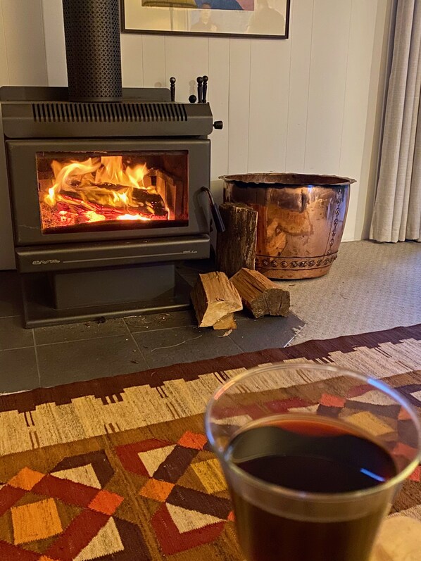 A wood-burning stove with logs, a copper pot, and a glass of red liquid on a patterned rug.