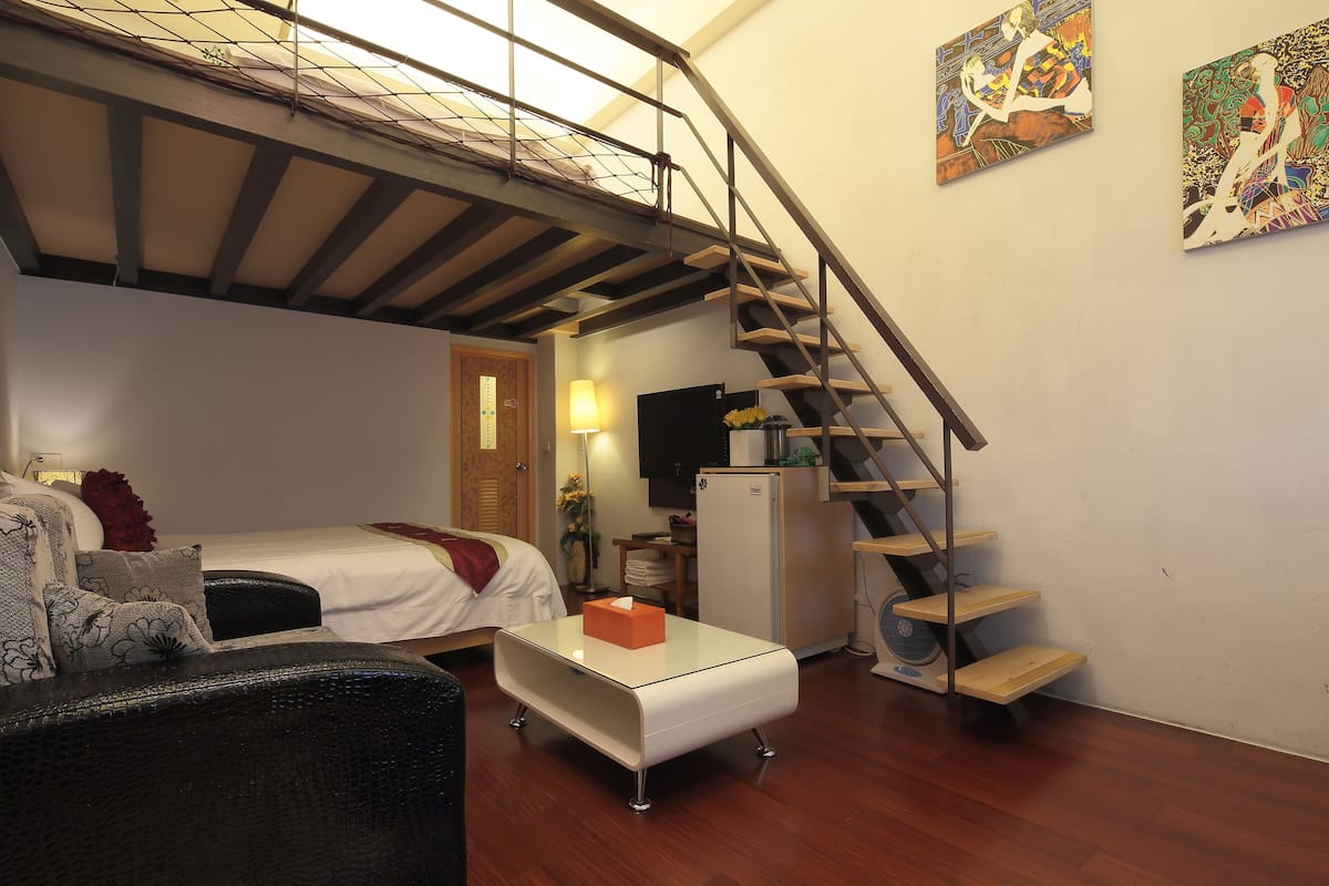 A compact living space with a bed, a sofa, a glass-top coffee table, a staircase, and two artworks on the walls.