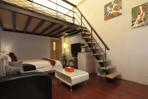 Un espace de vie compact comprenant un lit, un canapé, une table basse en verre, un escalier et deux œuvres d’art accrochées aux murs.