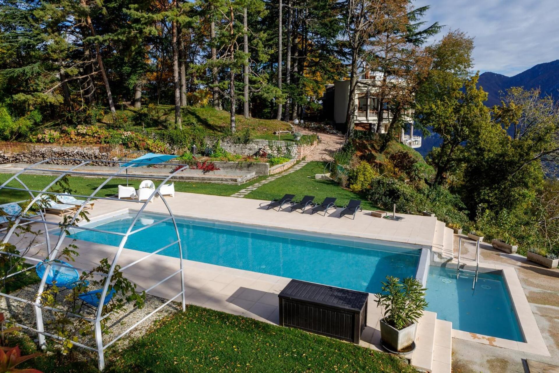Une piscine entourée de chaises longues, elle-même bordée d’un jardin et d’arbres, avec une vue sur les montagnes en arrière-plan.