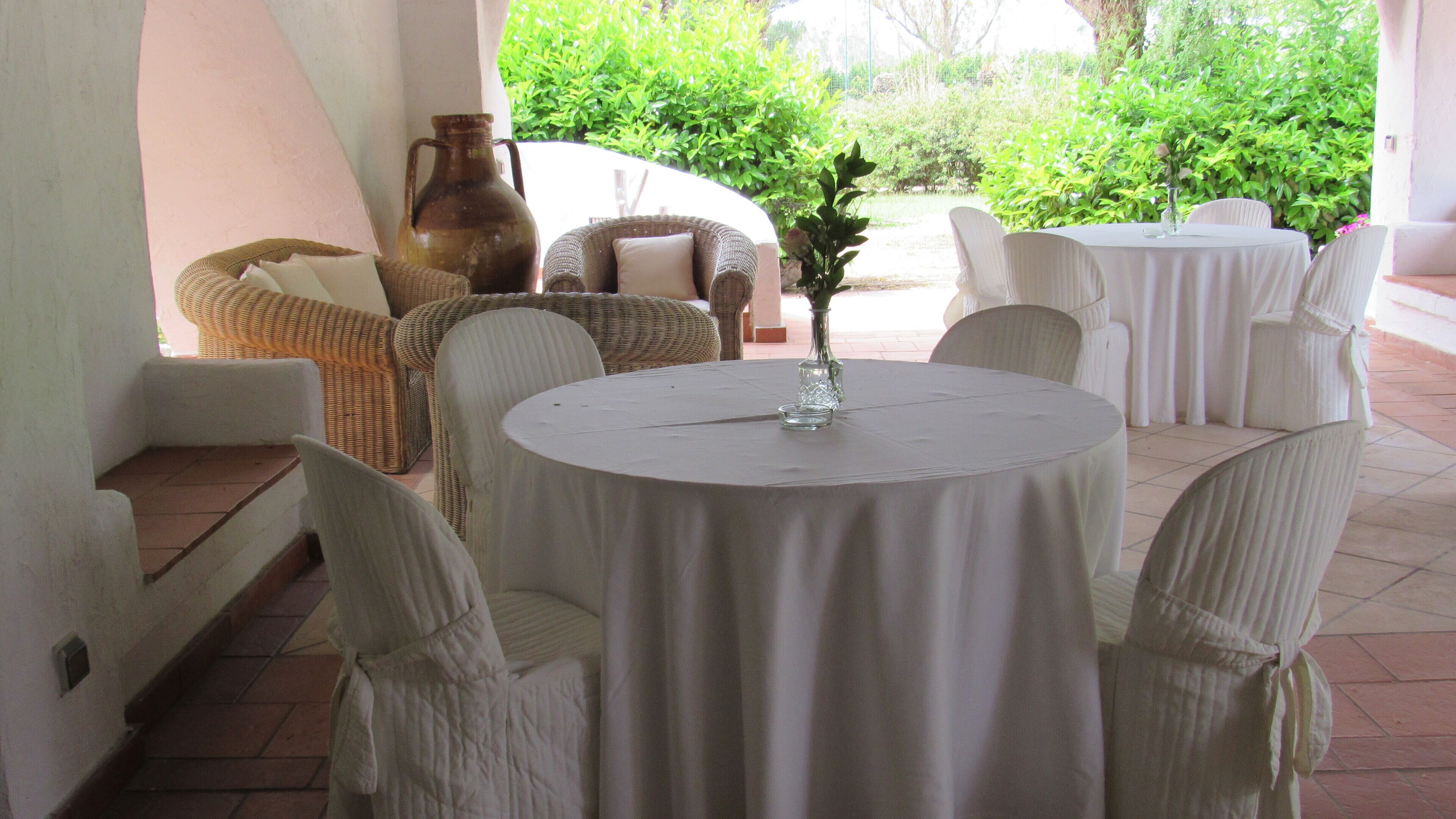A round table with a white tablecloth surrounded by white chairs, a vase with a flower, and wicker furniture in a covered outdoor area.