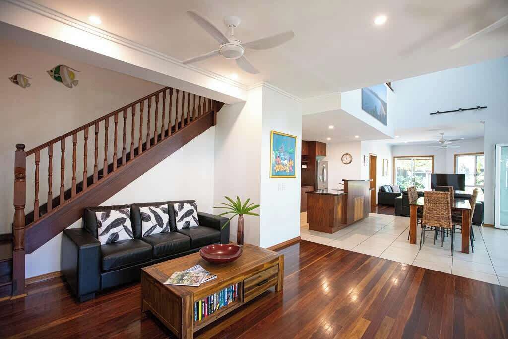 A spacious living area with a wooden staircase, a black sofa with patterned cushions, a coffee table with books, and a dining area with chairs and a table.