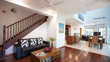 A spacious living area with a wooden staircase, a black sofa with patterned cushions, a coffee table with books, and a dining area with chairs and a table.