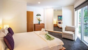 Una habitación de hotel con una cama grande, una zona de estar con una silla, un aparador de madera y un ventanal amplio.