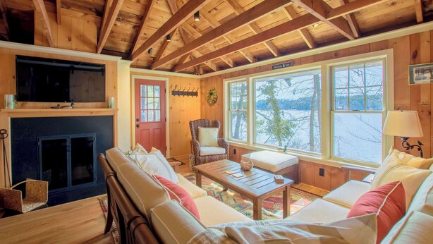 A cozy cabin interior with a fireplace, wooden beams, a sofa, a chair, and large windows offering a view of the outdoors. - Waterfront on Crystal Lake! (Orleans)