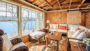 A cozy cabin interior with a large window offering a view of a lake, a wooden ceiling, and a sofa set. - Waterfront on Crystal Lake! (Orleans)