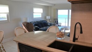A kitchen with a dishwasher, a sink, and a dining area with chairs. There is a view of the ocean through large windows. - Beachcomber unit 202 (Longboat Key)