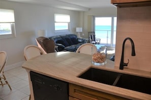 A kitchen with a dishwasher, a sink, and a dining area with chairs. There is a view of the ocean through large windows.