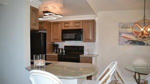 A modern kitchen with wooden cabinets, a black refrigerator, a microwave, and a stove. There is a dining area with a glass table and wicker chairs. - Beachcomber unit 202 (Longboat Key)
